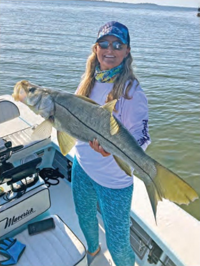 A smiling angler in a blue cap, sunglasses and a white long sleeve shirt holding up a big snook on a flats boat, with calm water behind her