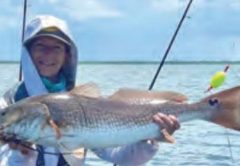 Wendy Biles in a white hooded sun shirt sitting on her pedal kayak and holding up a big redfish, with fishing rods, a popping cork and open water under a cloudy sky behind her