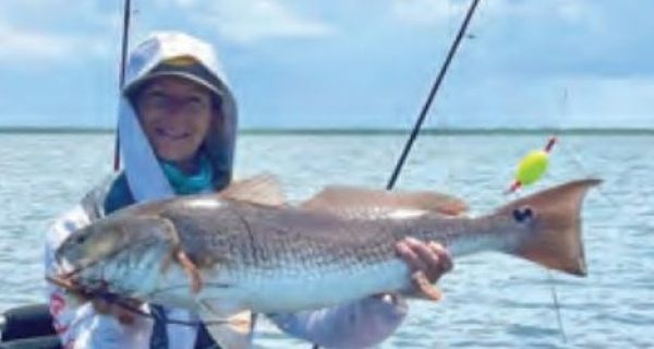 Wendy Biles in a white hooded sun shirt sitting on her pedal kayak and holding up a big redfish, with fishing rods, a popping cork and open water under a cloudy sky behind her
