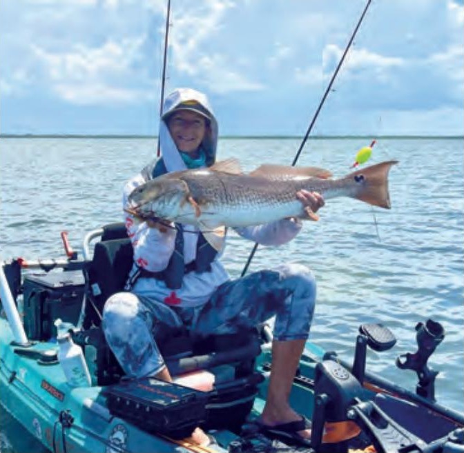 Wendy Biles in a white hooded sun shirt sitting on her pedal kayak and holding up a big redfish, with fishing rods, a popping cork and open water under a cloudy sky behind her