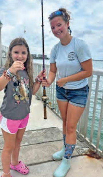A young girl in a gray shirt and pink shorts holding up a small fish on a fishing pier, next to a smiling young woman in a light blue T-shirt holding a fishing rod