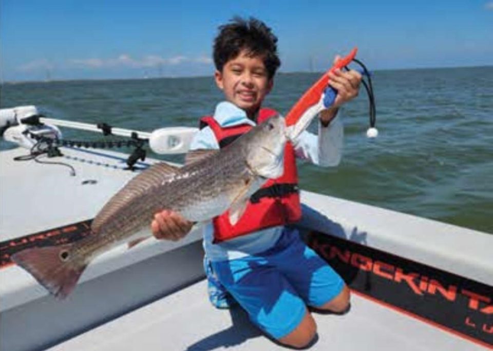 A smiling boy in a red life jacket kneeling on a boat deck and holding up a redfish with a fish grip, with open water behind him