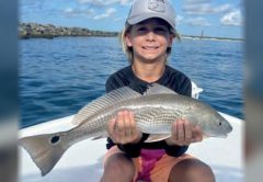 A smiling boy in a gray cap sitting on a boat and holding a redfish with both hands, with the Ponce Inlet jetty and lighthouse in the distance behind him