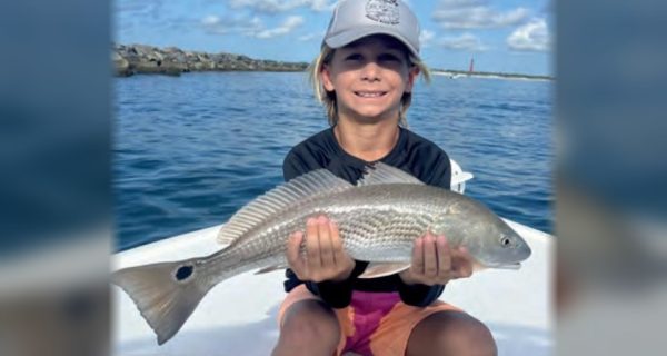 A smiling boy in a gray cap sitting on a boat and holding a redfish with both hands, with the Ponce Inlet jetty and lighthouse in the distance behind him