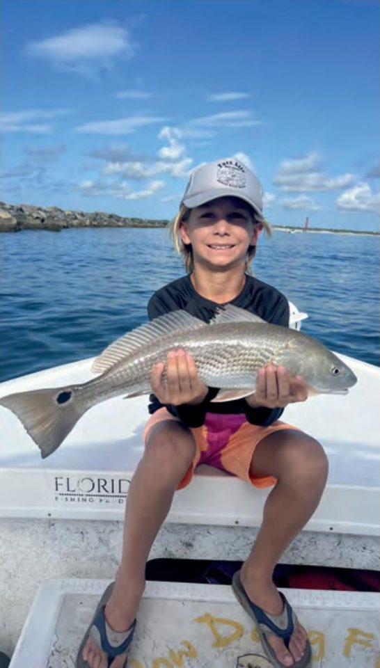 A smiling boy in a gray cap sitting on a boat and holding a redfish with both hands, with the Ponce Inlet jetty and lighthouse in the distance behind him