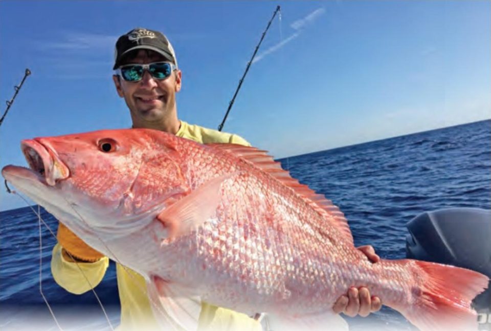 An angler in a black cap, mirrored sunglasses and a yellow long sleeve shirt holding up a big red snapper on a boat offshore, under a clear blue sky