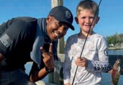 An Edgewater police officer in a black cap making a peace sign beside a smiling young angler in a white fishing shirt, who holds a rod with his small fish on the line, on a dock by the water