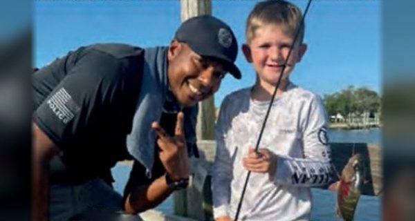 An Edgewater police officer in a black cap making a peace sign beside a smiling young angler in a white fishing shirt, who holds a rod with his small fish on the line, on a dock by the water