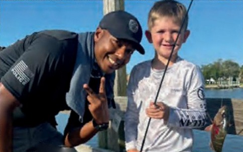 An Edgewater police officer in a black cap making a peace sign beside a smiling young angler in a white fishing shirt, who holds a rod with his small fish on the line, on a dock by the water