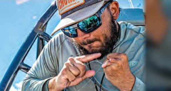 An angler in a cap, mirrored sunglasses and a gray long sleeve shirt tying a knot in fishing line on a boat, with a spool of Seaguar line on his knee