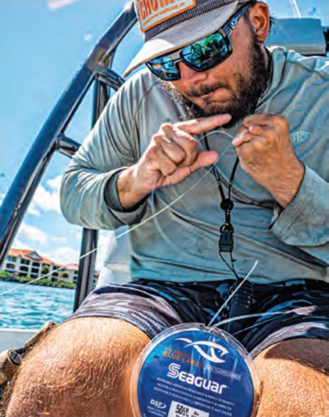 An angler in a cap, mirrored sunglasses and a gray long sleeve shirt tying a knot in fishing line on a boat, with a spool of Seaguar line on his knee