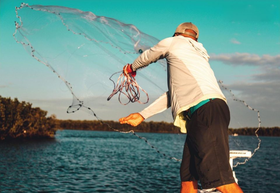 An angler in a cap and a long sleeve shirt throwing a cast net from the bow of a boat, the net spreading open against a teal sky over the water and a mangrove shoreline