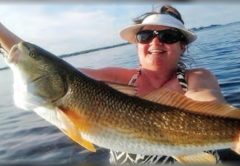 A smiling woman in a white visor and sunglasses holding up a big redfish on a boat, with a fishing rod and a purple float at left and calm water behind her