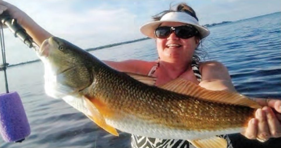 A smiling woman in a white visor and sunglasses holding up a big redfish on a boat, with a fishing rod and a purple float at left and calm water behind her