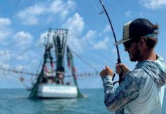 An angler in a black cap, sunglasses and a long sleeve fishing shirt fighting a fish with a bent rod from the bow of a boat, with a shrimp boat and a blue sky behind him