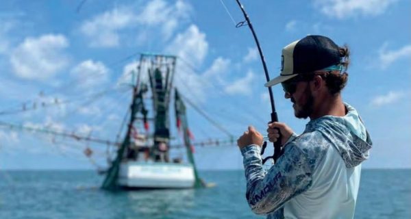 An angler in a black cap, sunglasses and a long sleeve fishing shirt fighting a fish with a bent rod from the bow of a boat, with a shrimp boat and a blue sky behind him