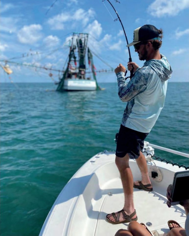 An angler in a black cap, sunglasses and a long sleeve fishing shirt fighting a fish with a bent rod from the bow of a boat, with a shrimp boat and a blue sky behind him