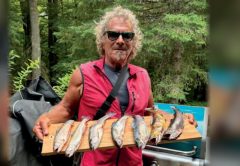 Marco Pompano in sunglasses and a red vest holding a wooden board lined with trout beside a grill, with a white dog looking up at him and trees behind