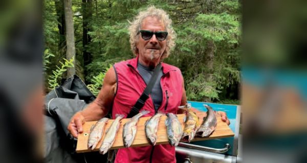 Marco Pompano in sunglasses and a red vest holding a wooden board lined with trout beside a grill, with a white dog looking up at him and trees behind