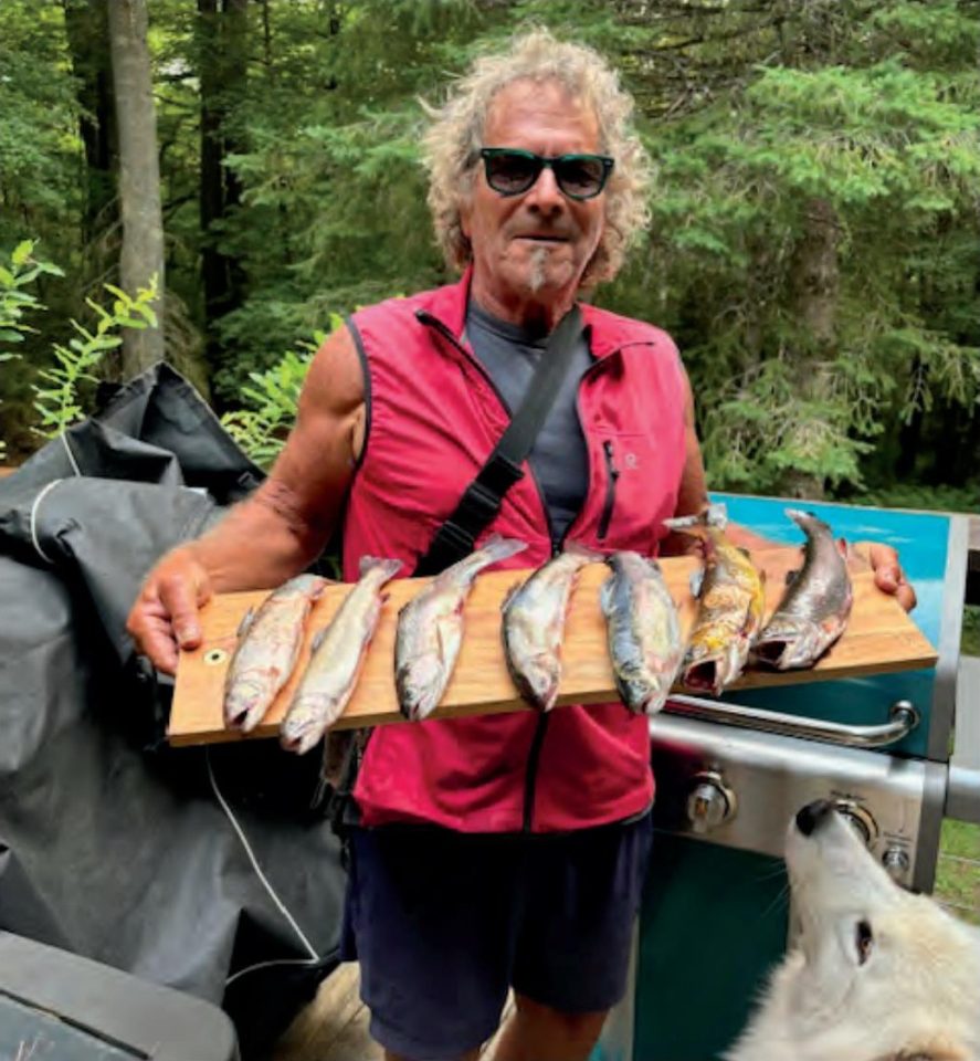 Marco Pompano in sunglasses and a red vest holding a wooden board lined with trout beside a grill, with a white dog looking up at him and trees behind