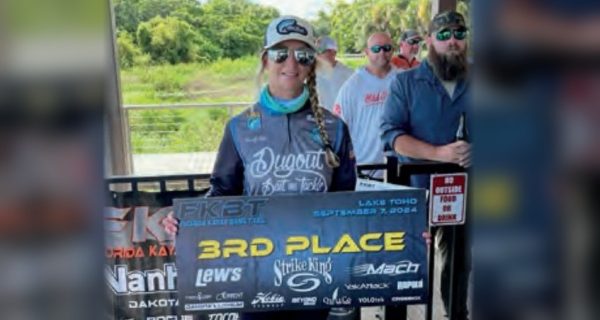 Wendy Biles in a cap, sunglasses and a tournament jersey holding a 3rd Place banner from the September 7, 2024 kayak bass tournament on Lake Toho, with other anglers behind her