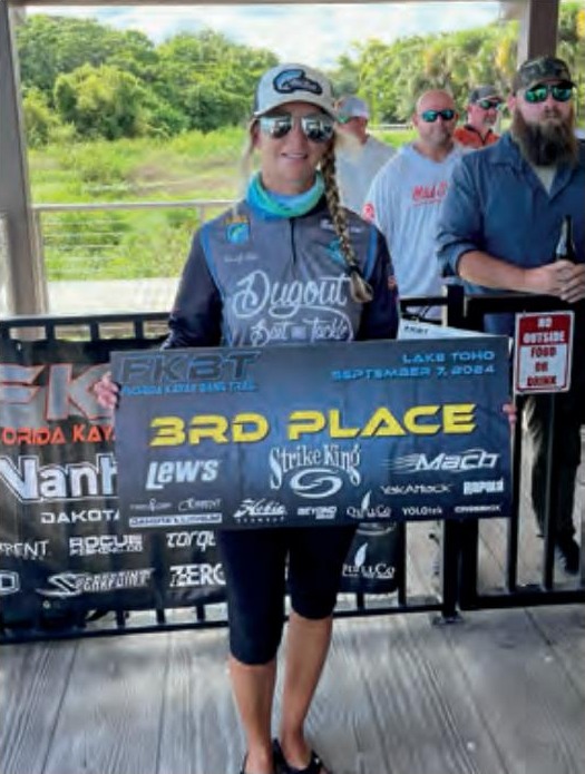 Wendy Biles in a cap, sunglasses and a tournament jersey holding a 3rd Place banner from the September 7, 2024 kayak bass tournament on Lake Toho, with other anglers behind her