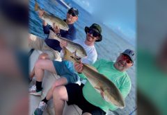 Three smiling anglers in caps and sunglasses sitting on a boat, each holding up a redfish, with blue water and another boat behind them