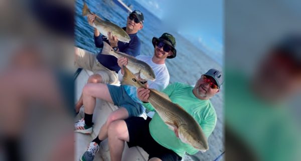 Three smiling anglers in caps and sunglasses sitting on a boat, each holding up a redfish, with blue water and another boat behind them