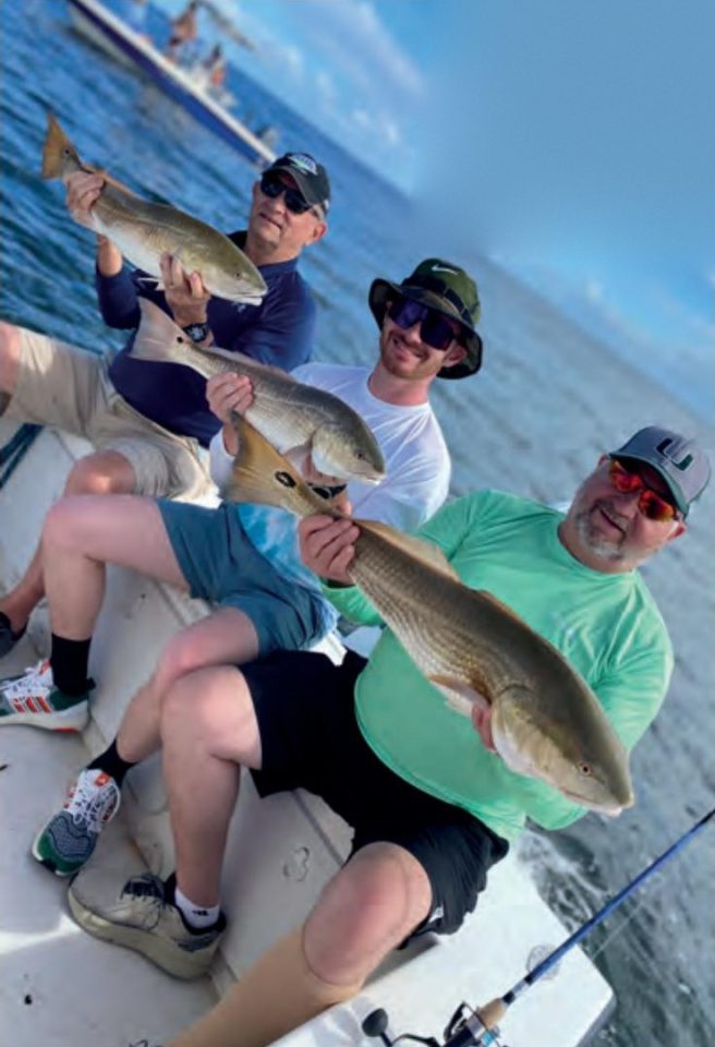 Three smiling anglers in caps and sunglasses sitting on a boat, each holding up a redfish, with blue water and another boat behind them