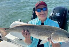 A smiling angler in a red cap, blue mirrored sunglasses and a light blue shirt sitting on a boat holding a big redfish, with an outboard motor and open water behind him