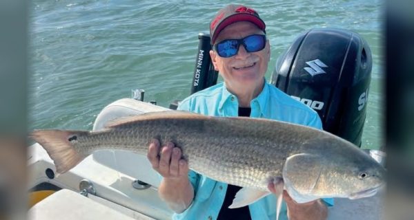 A smiling angler in a red cap, blue mirrored sunglasses and a light blue shirt sitting on a boat holding a big redfish, with an outboard motor and open water behind him