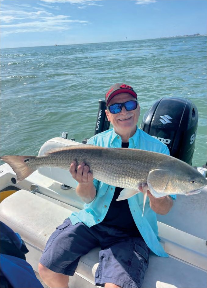 A smiling angler in a red cap, blue mirrored sunglasses and a light blue shirt sitting on a boat holding a big redfish, with an outboard motor and open water behind him