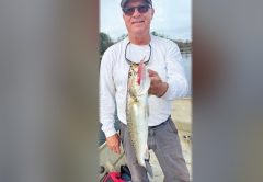 Tim Barefoot in a cap, sunglasses and a white long sleeve shirt holding up a spotted seatrout with a lure in its mouth, standing in a boat on the water