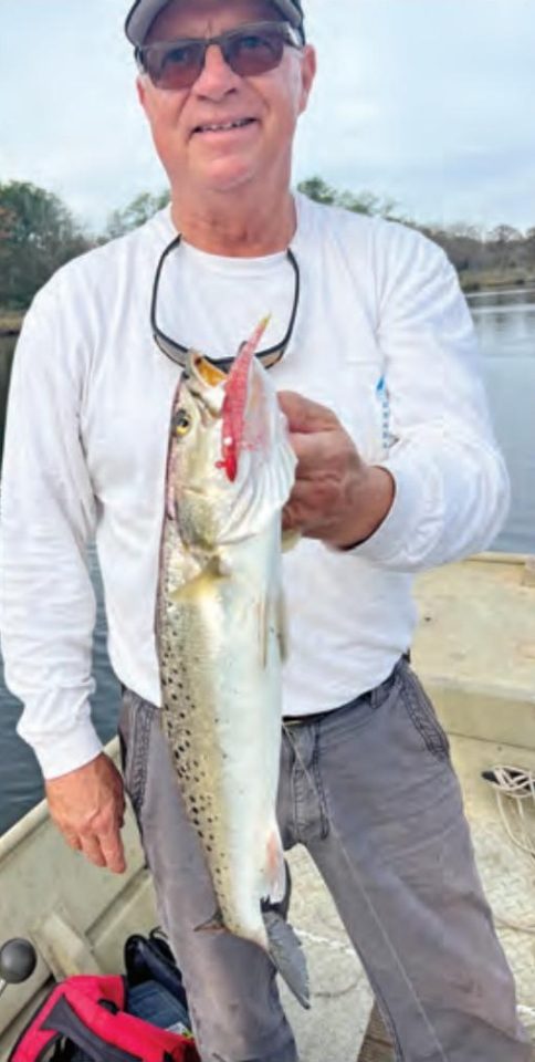 Tim Barefoot in a cap, sunglasses and a white long sleeve shirt holding up a spotted seatrout with a lure in its mouth, standing in a boat on the water