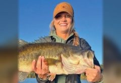 A smiling angler in an orange cap holding up a big largemouth bass with both hands under a clear blue sky