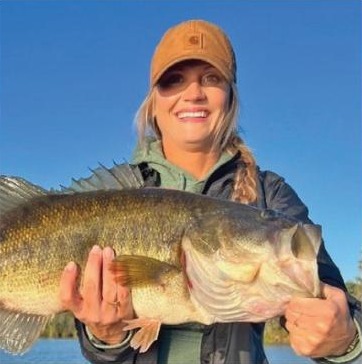 A smiling angler in an orange cap holding up a big largemouth bass with both hands under a clear blue sky