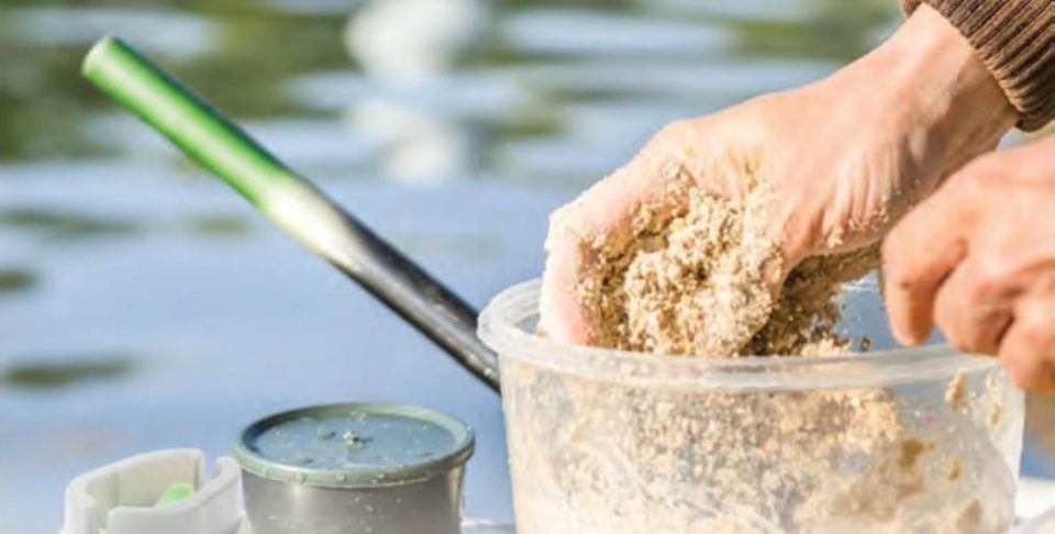 Hands mixing homemade chum in a clear plastic tub beside the water, next to a small bait container