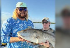 Capt. John Ellis in a mint green cap, sunglasses and a blue Reef Wrecker shirt holding up a big redfish on his boat beside an angler in a camouflage shirt and a colorful cap, with calm water and a tree line behind them