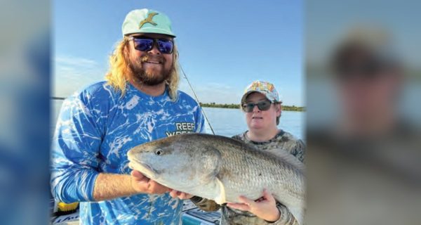 Capt. John Ellis in a mint green cap, sunglasses and a blue Reef Wrecker shirt holding up a big redfish on his boat beside an angler in a camouflage shirt and a colorful cap, with calm water and a tree line behind them