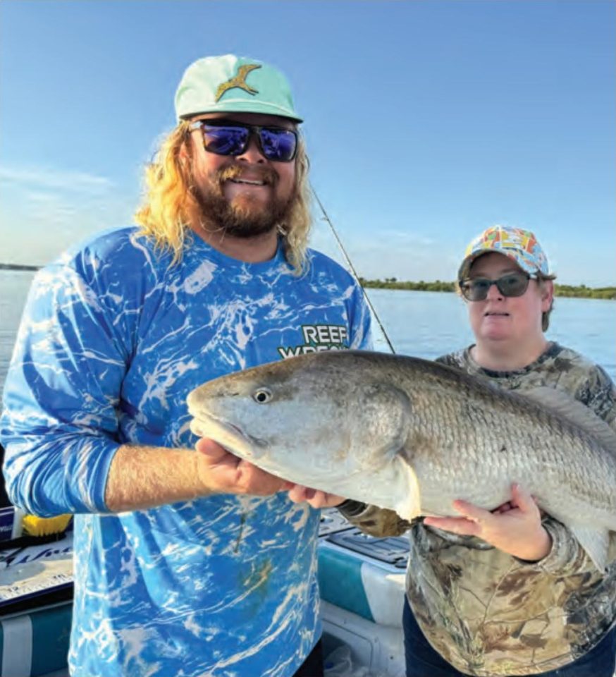 Capt. John Ellis in a mint green cap, sunglasses and a blue Reef Wrecker shirt holding up a big redfish on his boat beside an angler in a camouflage shirt and a colorful cap, with calm water and a tree line behind them