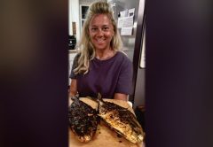 A smiling woman in a kitchen holding a wooden board with two whole pompano grilled with Chinese chili crisp