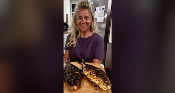 A smiling woman in a kitchen holding a wooden board with two whole pompano grilled with Chinese chili crisp