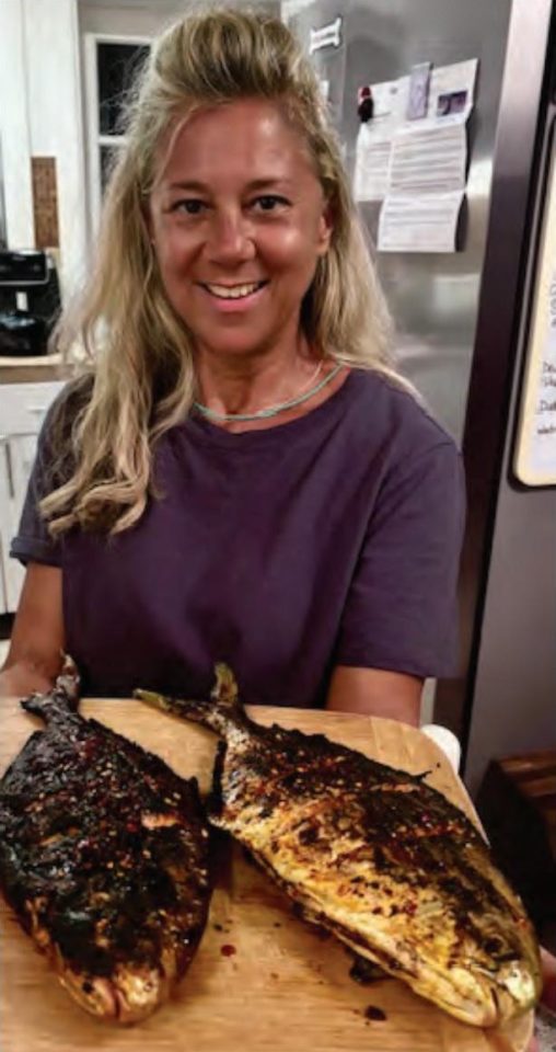 A smiling woman in a kitchen holding a wooden board with two whole pompano grilled with Chinese chili crisp