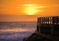 The sun glowing through an orange sky over the ocean, with waves breaking on the rocks below a railed pier