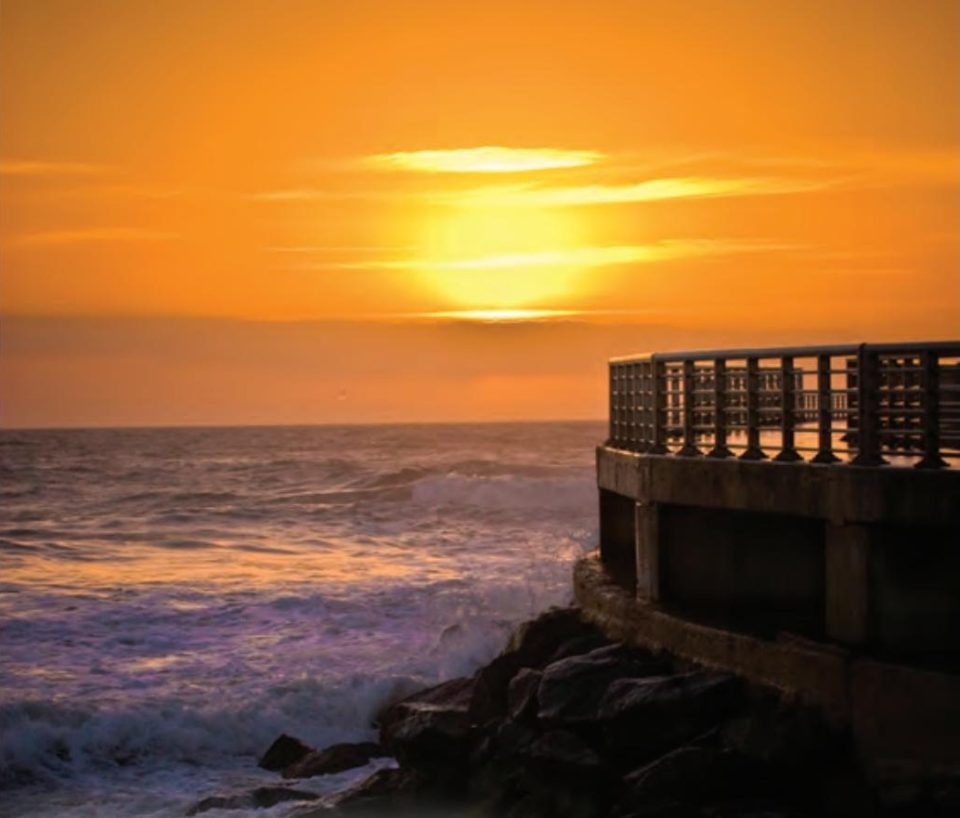The sun glowing through an orange sky over the ocean, with waves breaking on the rocks below a railed pier
