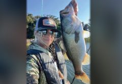 Wendy Biles in a trucker cap, mirrored sunglasses and a camouflage jacket taking a selfie while holding up a big largemouth bass, with trees and blue sky behind her