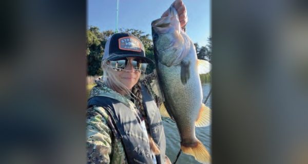 Wendy Biles in a trucker cap, mirrored sunglasses and a camouflage jacket taking a selfie while holding up a big largemouth bass, with trees and blue sky behind her
