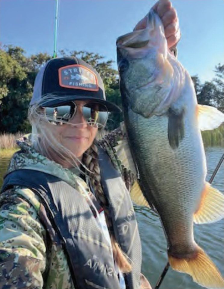 Wendy Biles in a trucker cap, mirrored sunglasses and a camouflage jacket taking a selfie while holding up a big largemouth bass, with trees and blue sky behind her