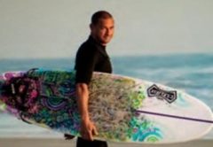 Jorge Alvarado in a black wetsuit walking barefoot along the beach with a colorful surfboard under his arm, with the ocean behind him