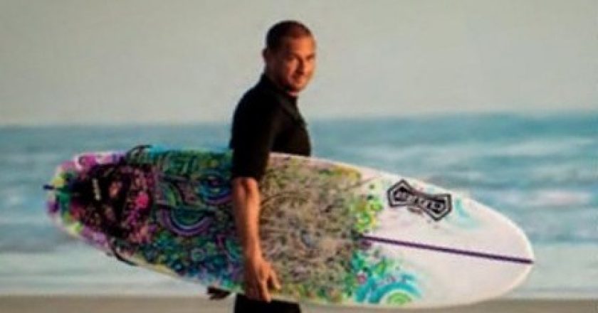 Jorge Alvarado in a black wetsuit walking barefoot along the beach with a colorful surfboard under his arm, with the ocean behind him